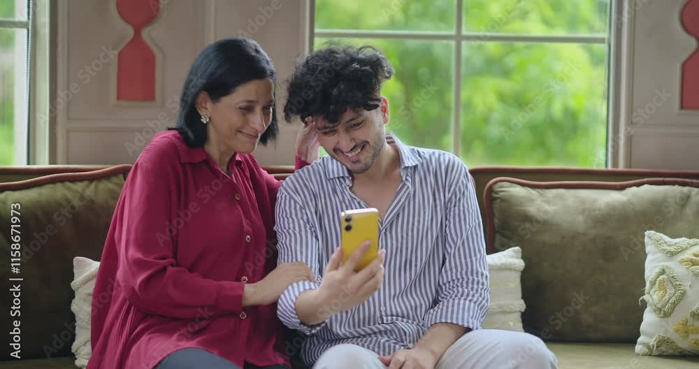 Indian Asian happy older woman mom lady young adult man male son sitting cozy sofa couch use mobile phone talk video call make funny gossip look camera joy enjoy fun day spend time indoor house