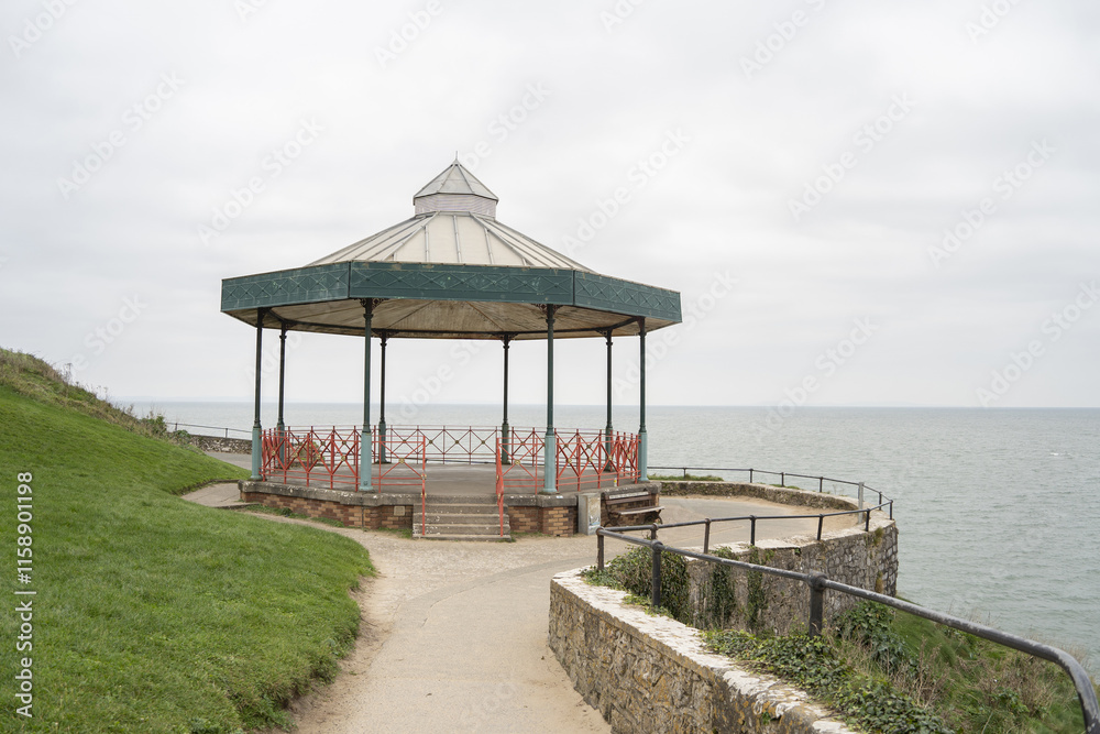 Fototapeta premium The image features a charming seaside bandstand made of metal and wood, situated on a grassy coastal cliff overlooking the ocean. The structure is octagonal with a peaked roof.