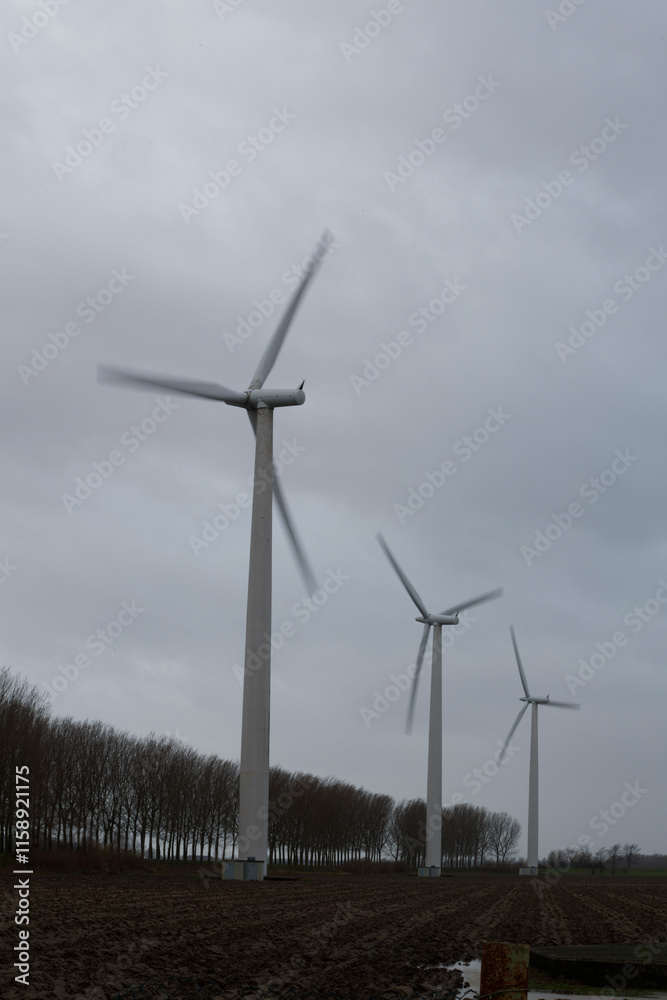 Wind turbines stand in a plowed field, amidst bare trees under a cloudy sky. Renewable energy, rural landscape. Wind turbines stand in a plowed field, amidst bare trees under a cloudy sky. Renewable energy, rural landscape.