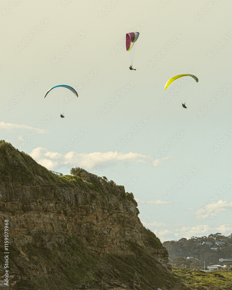 Three colorful paragliders gracefully glide above a lush coastal cliff, bathed in soft, warm light. The image captures a sense of freedom and adventure against the backdrop of a clear sky. The vibrant Three colorful paragliders gracefully glide above a lush coastal cliff, bathed in soft, warm light. The image captures a sense of freedom and adventure against the backdrop of a clear sky. The vibrant