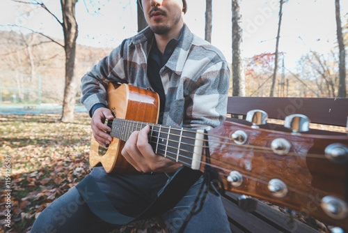 Man playing acoustic guitar outdoors in autumn, wearing a plaid shirt, sitting on a wooden bench surrounded by fallen leaves. Perfect for themes of music, relaxation, and nature photo