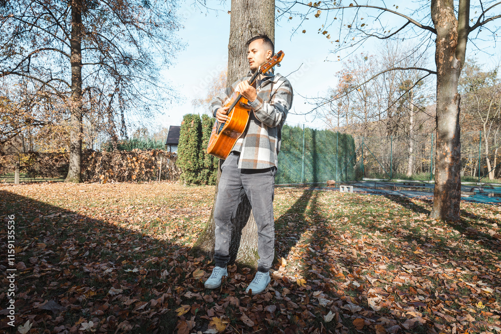Young man leaning against a tree, playing an acoustic guitar in a sunlit autumn park, surrounded by fallen leaves and nature’s vibrant colors, creating a serene atmosphere