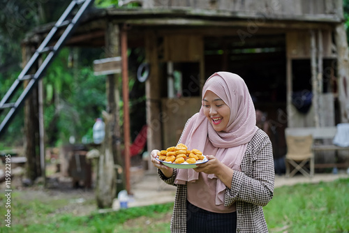 young Indonesian woman in hijab standing and posing with a plate full of nuggets in front of a forest cabin, travel concept. photo