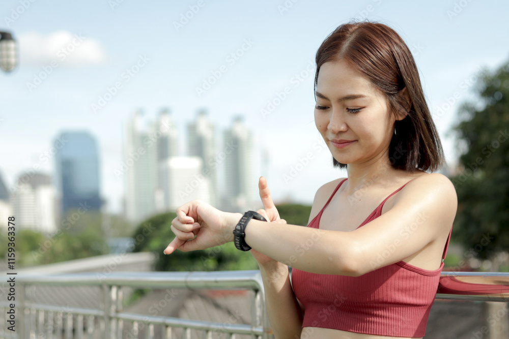 Fototapeta premium Short haired sporty girl in sportswear looks and touches smartwatch after jogging work out for exercise on city street. Happy smiling Asian woman do sport fitness outside. Healthy outdoor activity.
