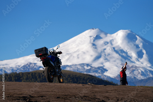 Traveling by bike along the mountain roads of the Caucasus. A bike against the background of snowy Elbrus and blue sky. A biker with a phone in his outstretched hand. Copy space photo