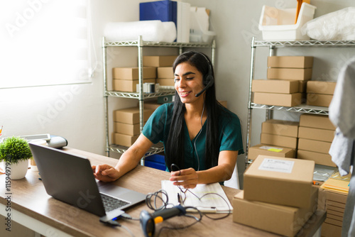 Hispanic business owner wearing a headset while overseeing online orders and deliveries in her warehouse with a laptop photo