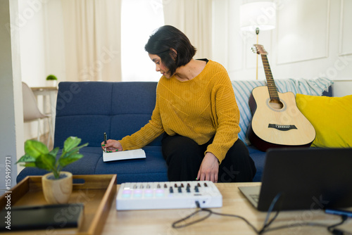 Hispanic musician relaxing on home studio sofa, strumming acoustic guitar and composing melodic musical notes with creative passion