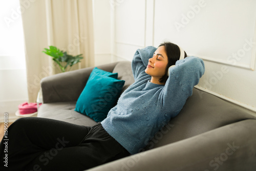 Peaceful female relaxing on soft couch, listening to music through headphones, eyes closed, hands resting behind head