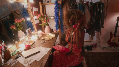 Full lowkey shot of 70s young Black female singer with puffy hair in open dress sitting at table in dressing room, smiling, laughing and joyfully sharing her impressions, talking on red phone photo