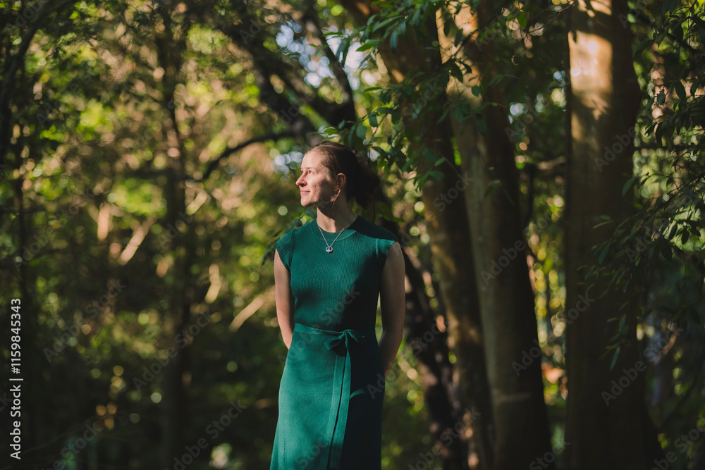 A woman in a green dress stands amidst tall trees in soft sunlight, her smile radiating warmth and her surroundings rich with forest tones and depth A woman in a green dress stands amidst tall trees in soft sunlight, her smile radiating warmth and her surroundings rich with forest tones and depth