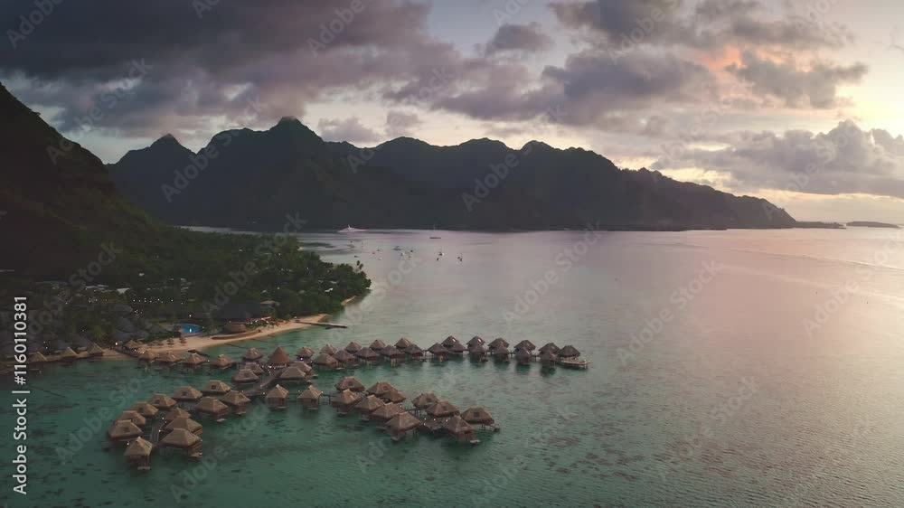 Aerial view of luxury overwater bungalows in turquoise lagoon at sunset reflecting clouds and dramatic sky with lush mountains in background in Moorea, French Polynesia. Remote wild nature paradise