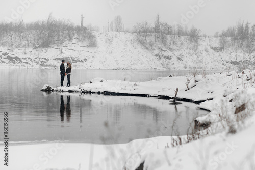 winter scene by the pond. In the foreground you can see two people standing on the snowy shore. They hold hands and look at each other. There's snow all around them photo