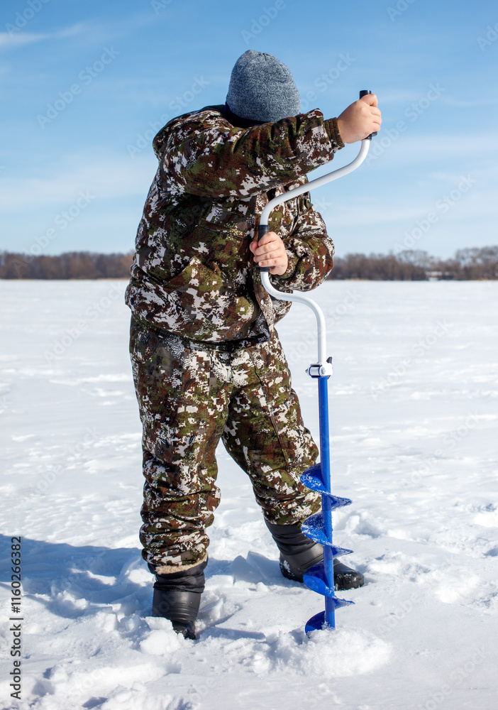 A man in camouflage clothing is standing in the snow with a blue ice auger A man in camouflage clothing is standing in the snow with a blue ice auger