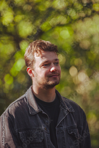 A young man wearing a denim jacket with a backpack stands confidently on a shaded pathway surrounded by lush greenery, enjoying the serene environment of a park