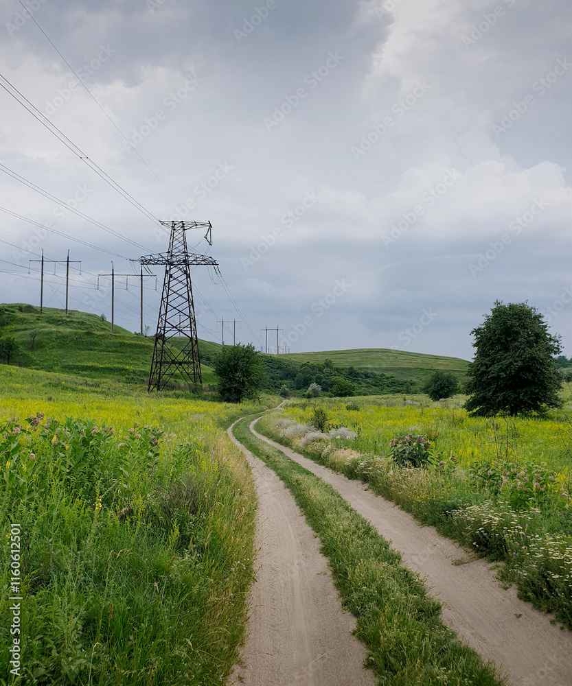 Dirt road amid hilly terrain, few transmission towers on top of the hills. Curvy trail amid meadow in a small valley, cloudy sky in the background. Dirt road amid hilly terrain, few transmission towers on top of the hills. Curvy trail amid meadow in a small valley, cloudy sky in the background.