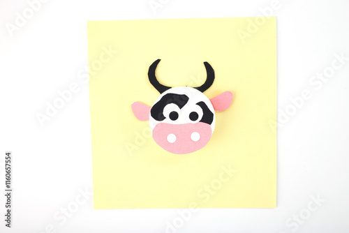 Handmade cow craft made from a white bottle cap with black spots, pink paper muzzle, and ears on a yellow square background. Flat lay composition with copy space. DIY and kids craft concept, top view photo