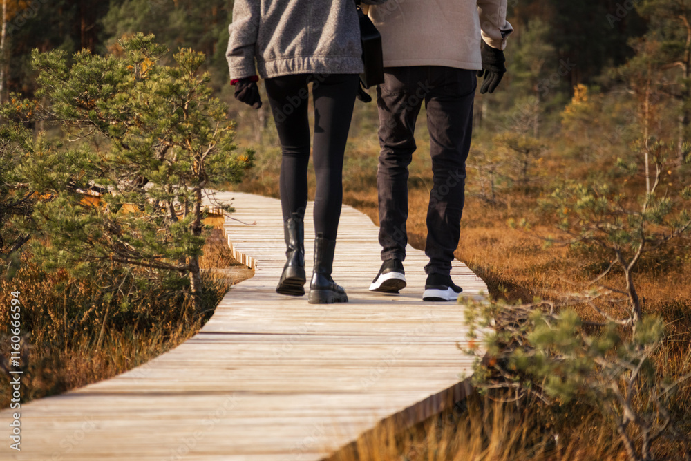 People are walking on a wooden boardwalk in the forest. The woman is dressed in dark pants and boots, and the man in dark pants and sneakers.
People are walking on a wooden boardwalk in the forest. The woman is dressed in dark pants and boots, and the man in dark pants and sneakers.