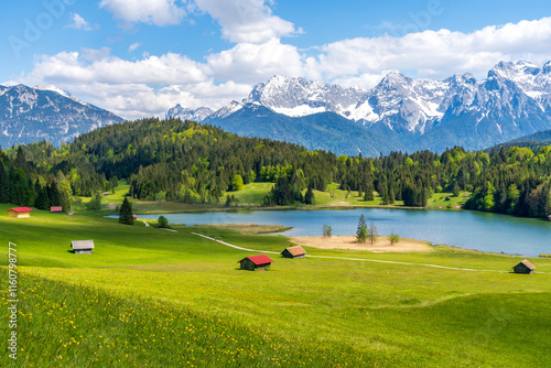 Geroldsee lake in Bavaria, Germany, featuring rustic wooden huts and the majestic, snow-dusted Karwendel mountains under a sunny blue summer sky photo