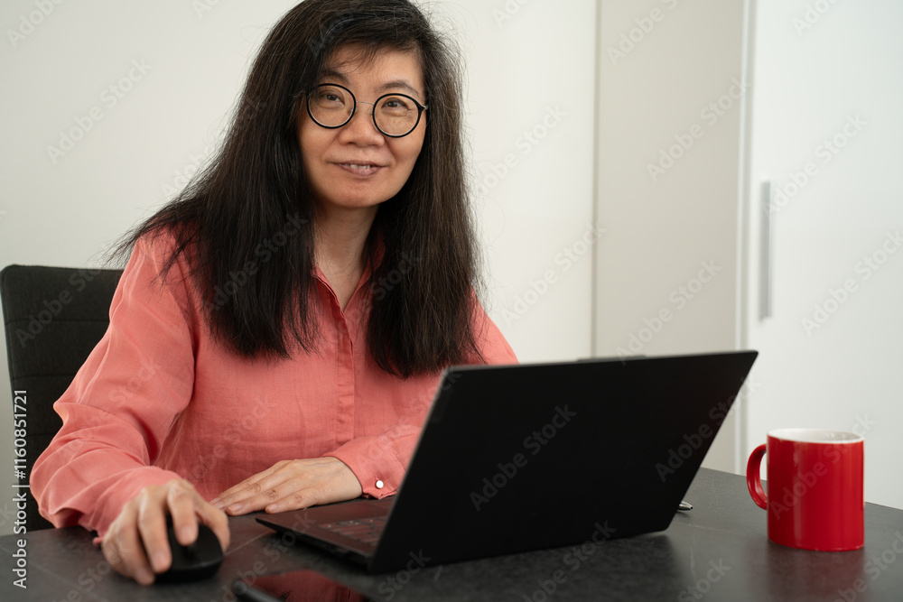 Naklejka premium Middle aged chinese business woman with glasses, long black hair and red blouse is working with a laptop inside an office, just looking up from her work. Red cup of coffee near by.