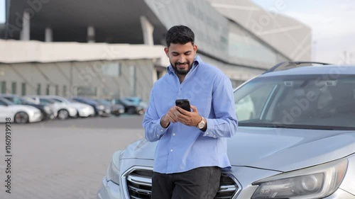 Attractive stylish business man checking smartphone, while leaning on grey car on parking lot. Front view of smiling bearded guy holding cell phone, messaging with friends. Concept of lifestyle.