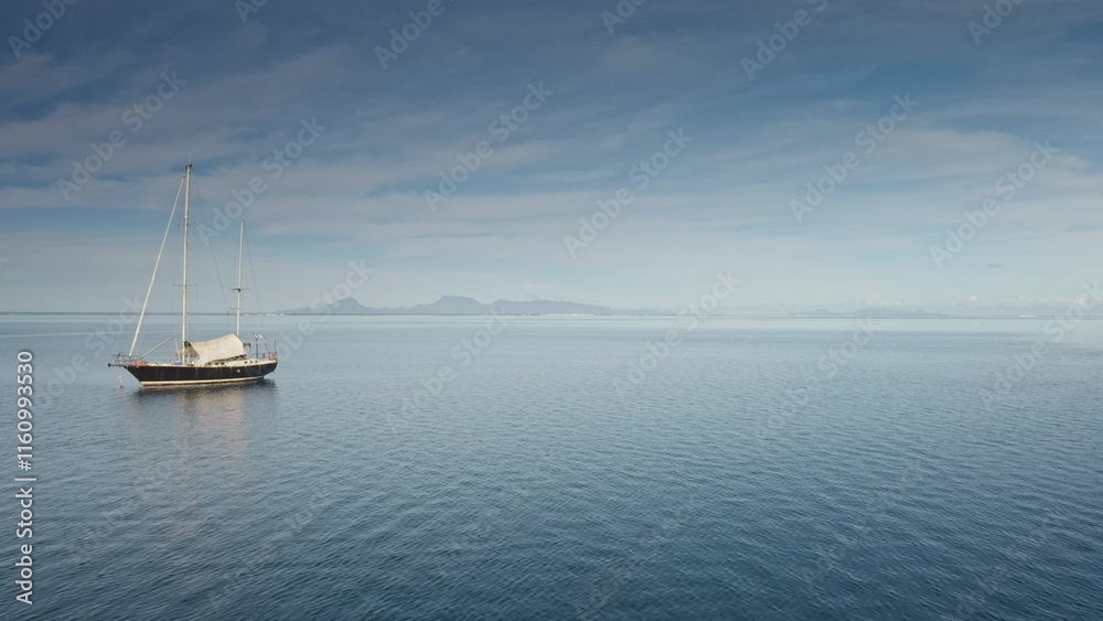 Fototapeta premium Wooden yacht anchor peacefully on serene tropical sea, with distant island visible on horizon under cloudy blue sky. Remote wild nature paradise, exotic summer travel destination. Drone flight