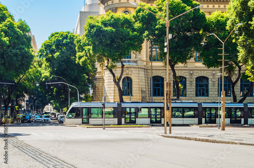 A modern tram travels along the street in Rio de Janeiro, surrounded by lush green trees and historic buildings.