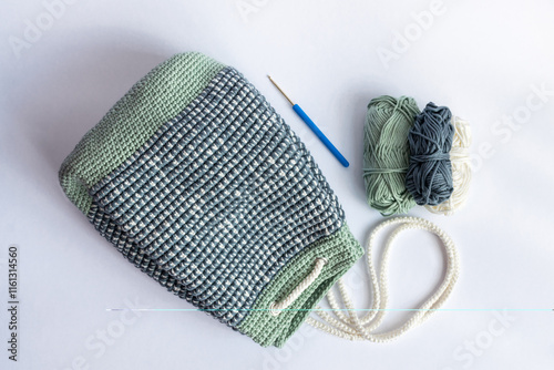 Handmade Crocheted Mosaic Bucket Bag Backpack in Pastel green, White and Gray colors on white background with crochet hook and three balls of yarn photo