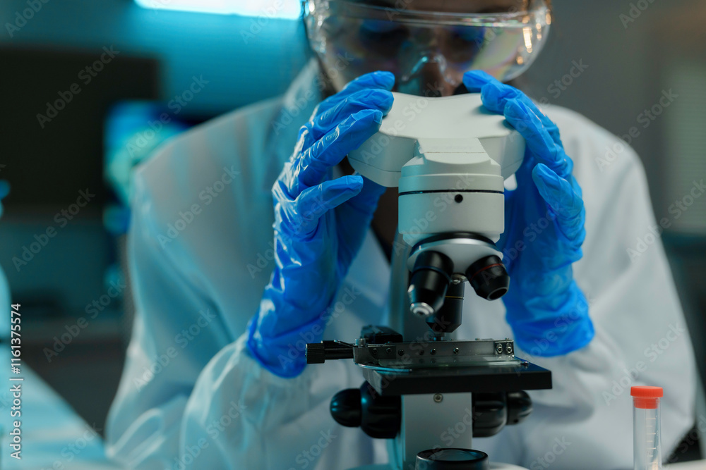 Scientist wearing protective glasses and gloves using microscope examining samples in a laboratory