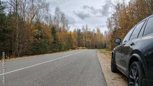 A short stop during a autumn road trip. Car parked on side of paved highway with markings, sand shoulders and fallen leaves. A mixed forest stands on the sides. There is yellow foliage on the trees