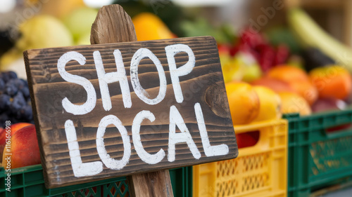 A rustic wooden sign reading 'Shop Local' stands prominently among vibrant fruits at a farmer's market, promoting community support and sustainability. photo