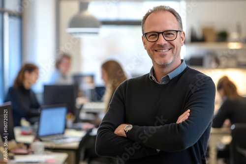 A confident businessman in his office, standing with arms crossed and smiling at the camera while people work behind him. photo