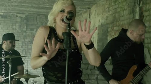 Female singer in black clothes and dark makeup passionately singing into a microphone with arm raised, while bandmates create powerful rock music in an industrial setting photo