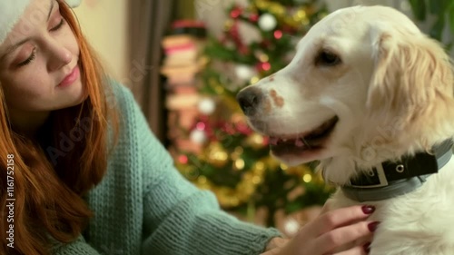 A young woman wearing a Santa hat cuddles her golden retriever on a cozy cushion. The festive Christmas tree in the background creates a heartwarming holiday atmosphere