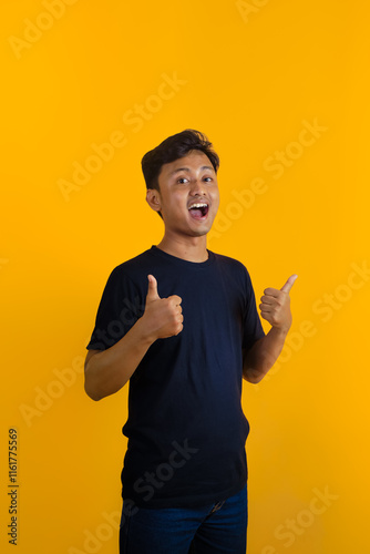 A young Asian man in a black shirt gives a thumbs up and smiles confidently against a dark yellow background. This image depicts positivity, approval, and satisfaction.