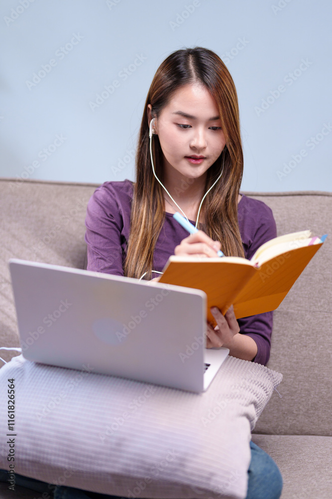 A woman sitting on a sofa with earphones, taking notes while attending an online class, using a laptop and a notebook A woman sitting on a sofa with earphones, taking notes while attending an online class, using a laptop and a notebook