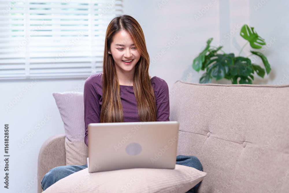 A young woman working on a laptop while sitting on a sofa, smiling and appearing focused, in a cozy home setting A young woman working on a laptop while sitting on a sofa, smiling and appearing focused, in a cozy home setting