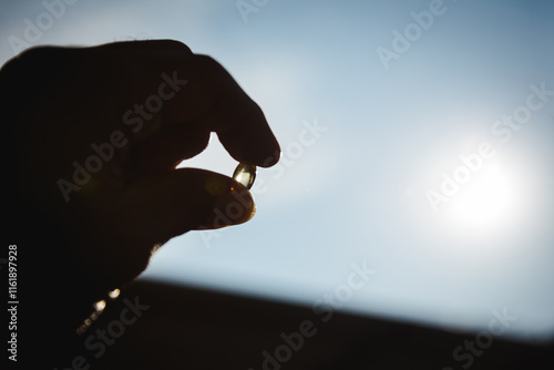Image of a hand holding a shiny yellow vitamin pill against a sunny orange-golden sky in the morning and evening.