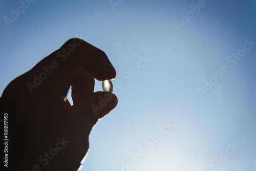 Image of a hand holding a shiny yellow vitamin pill against a sunny orange-golden sky in the morning and evening.