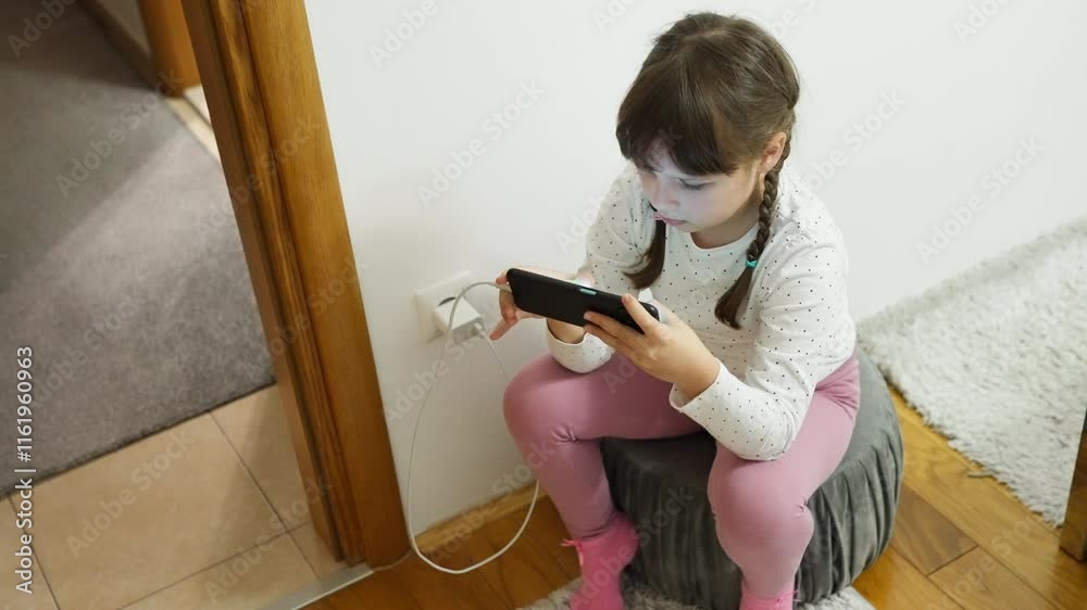 Young girl sitting indoors, focused on a smartphone while it charges. Concept of childhood technology use, screen time, and phone addiction
