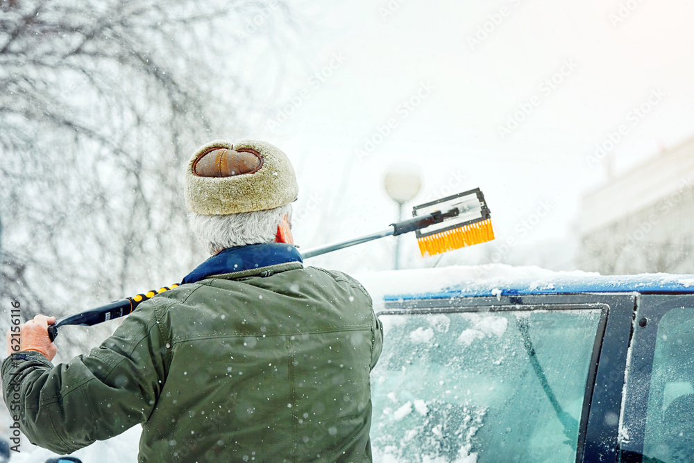 Man sweeps snow off the rood of car during heavy snowfall in an icy parking area. Senior man cleans snow covered car using brush amidst snowfall and icy conditions, snowy day struggles.