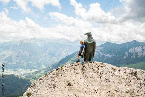 13 july 2024, Roda di Vael, Italy. Caucasian hiker standing at the Christomannos monument, a bronze eagle in front of the  mighty walls of the Catinaccio-Rosengarten massif Italian Dolomites, hiking d photo