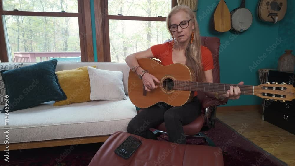 A middle-aged woman is seated in a comfortable chair, strumming acoustic guitar while focusing on her music. The room features instrumental decor, creating a warm atmosphere.