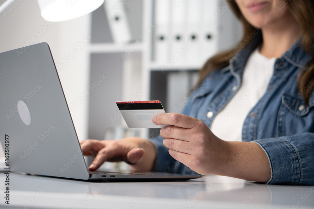 Fototapeta premium A young woman engages in online shopping, actively using her credit card while sitting at her desk with a laptop. This modern consumer experience exemplifies convenience and the digital lifestyle