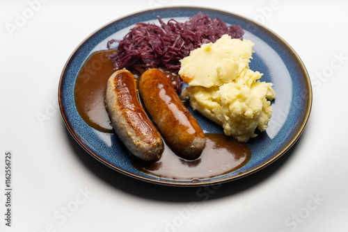 , Deutschland, 29.12.2024,  Bratwürste mit dunkler Soße, Rotkohl und cremigem Kartoffelpüree auf einem blauen Teller – ein klassisches deutsches Gericht.