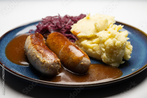, Deutschland, 29.12.2024,  Bratwürste mit dunkler Soße, Rotkohl und cremigem Kartoffelpüree auf einem blauen Teller – ein klassisches deutsches Gericht.