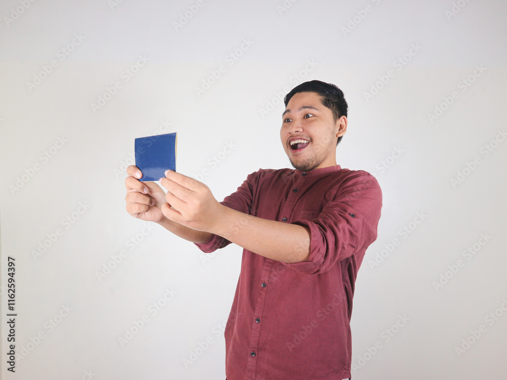 Smiling Asian man in a red shirt looks at the passport he is holding with an excited face Smiling Asian man in a red shirt looks at the passport he is holding with an excited face