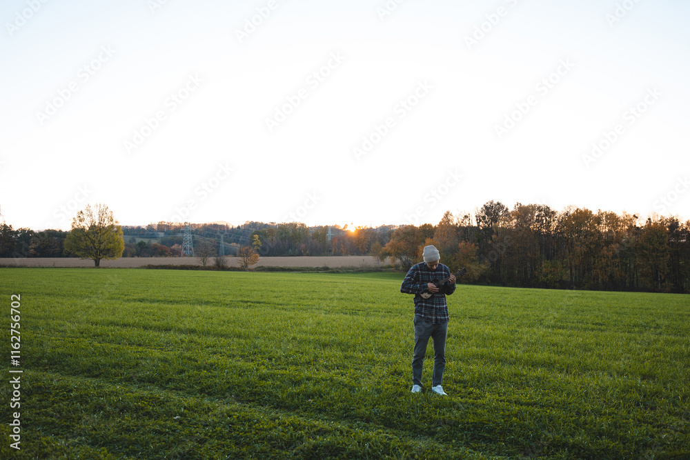 Musician in a plaid shirt and beanie plays the ukulele in a green field at sunset. The serene rural scene beautifully combines music, nature, and tranquility Musician in a plaid shirt and beanie plays the ukulele in a green field at sunset. The serene rural scene beautifully combines music, nature, and tranquility