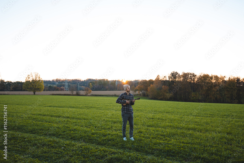 Musician in a plaid shirt and beanie plays the ukulele in a green field at sunset. The serene rural scene beautifully combines music, nature, and tranquility Musician in a plaid shirt and beanie plays the ukulele in a green field at sunset. The serene rural scene beautifully combines music, nature, and tranquility