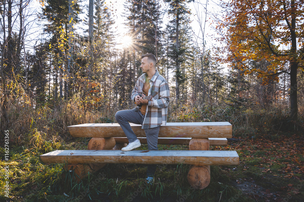 Fototapeta premium Musician stands by a rustic wooden bench in an autumn forest, playing the ukulele. With colorful leaves, a gazebo in the background, and a serene atmosphere, the scene reflects a peaceful connection
