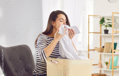 Woman smells clothes after using a new fabric conditioner. Young girl sitting on an armchair with a laundry basket and enjoying a nice, fresh, natural aroma of a clean white office shirt in her hands